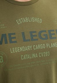 Olivovo zelené bavlnené tričko s tmavozeleným textom. Text obsahuje "Založené," "JA LEGEND," a podrobnosti o nákladnom lietadle Catalina CV283.