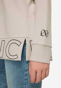 Sweatshirt beige avec des poignets côtelés, des fentes latérales et un texte noir audacieux au bas. Présente un logo graphique sur la manche. Fabriqué en tissu doux.