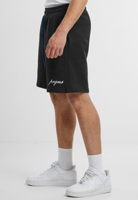 Mustat puuvillashortsit, joissa on joustava vyötärö ja sininen nyöri. Vasemmassa lahkeessa on valkoiset brodeeratut tekstit "pequs". Valkoisia tennareita käytetään.