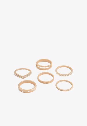 Six gold-tone rings, some with embedded clear stones, arranged on a white background, featuring varied textures and patterns.
