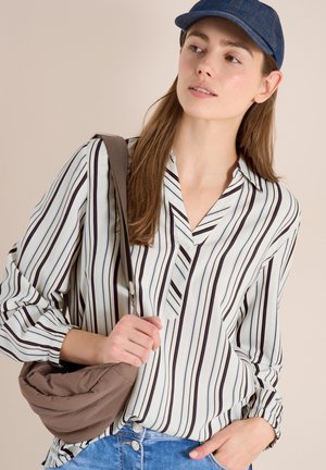 Giovane donna con berretto blu, camicetta a righe bianche e nere, che tiene una borsa marrone a tracolla e indossa jeans blu su uno sfondo neutro.