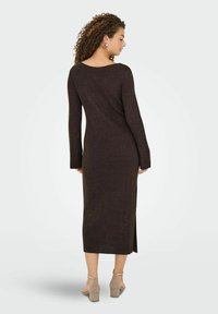 Kvinde med krøllet hår iført en lang, mørk rippet strikkjole og beige blokhæl sandaler, vendt væk med et sideprofil synligt.
