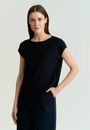 Junge Frau mit langen braunen Haaren, die ein schwarzes kurzärmeliges Oberteil und dunkle Hosen trägt, mit einer Hand in der Tasche vor hellem Hintergrund stehend.