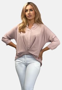 Jasnoróżowa bluzka o luźnym kroju z dekoltem w serek, rękawami do łokcia i skręconym detalem u dołu, zestawiona z dopasowanymi białymi spodniami.