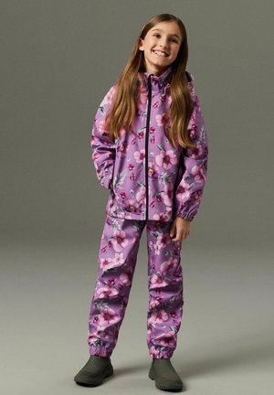 Dekle, ki se smeji, z rokami v žepih, oblečeno v vijolično dežno jakno z rožnim vzorcem in ujemajoče se hlače, stoji pred enobarvnim ozadjem.