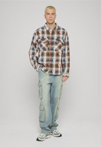 Camicia a quadri marrone e bianca con bottoni e due tasche sul petto, abbinata a pantaloni cargo azzurri chiaro con tasche laterali e vestibilità ampia.