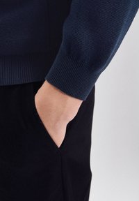 Donker marineblauwe gebreide trui met geribbelde manchetten, gecombineerd met zwarte broek. Close-up toont een hand in de zak, benadrukt de stoftextuur en pasvorm.