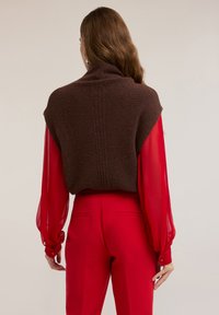 Osoba o długich brązowych włosach ubrana w prześwitującą czerwoną bluzkę, czerwone spodnie z wysokim stanem oraz bezrękawnik z ciemnobrązowej wełny, pokazana od tyłu.