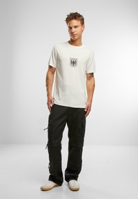 Vit bomull t-shirt med en svart emblem på bröstet, tillsammans med svarta cargobyxor och vita sneakers med bruna sulor.