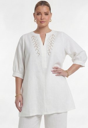 Kvinde iført hvid linned tunika med trekvartlange ærmer og guldbroderi med perler nær halsudskæringen, kombineret med matchende hvide bukser.