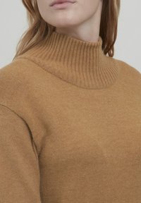Pulover bej cu guler înalt, fabricat dintr-un material tricotat moale, având un guler cu nervuri și o textură netedă, cu detalii de coasere minime.