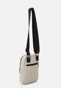 Beige leather crossbody bag with a textured surface, black adjustable strap, zip closure, and a front pocket with a black zipper.