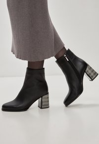Botas de cuero negro hasta el tobillo con forma afilada, que cuentan con un tacón de bloque con diseño metálico y una cremallera lateral para el cierre.