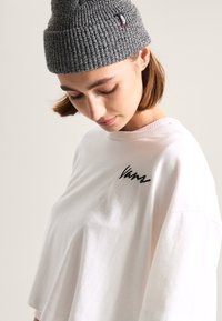 Camiseta de manga corta color rosa claro con un logo bordado en negro. La camiseta se acompaña de un gorro de lana gris con un pequeño parche. Textura de algodón suave.