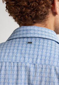 Camicia a motivi geometrici blu con colletto prominente, tessuto testurizzato e una piccola etichetta del marchio vicino al collo nella parte posteriore.
