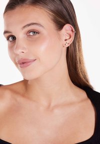 Modèle portant plusieurs boucles d'oreilles ; en métal doré et argenté, designs circulaires et en puce, sur un fond clair. Peau lisse, haut neutre.