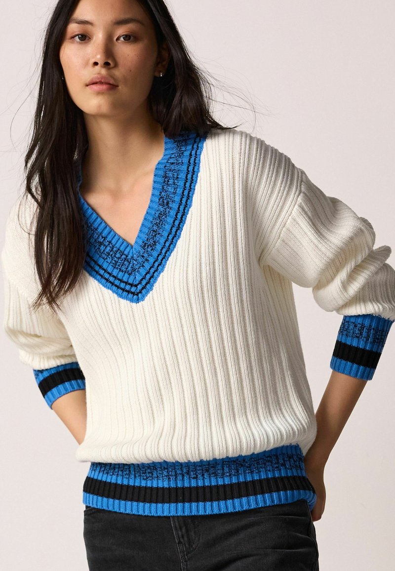 Weißer gerippter Pullover mit tiefblauem V-Ausschnitt und Detail am Hals. Blau- und schwarzgestreifte Akzente an den Ärmelbündchen und am Saum. Weiche Textur.