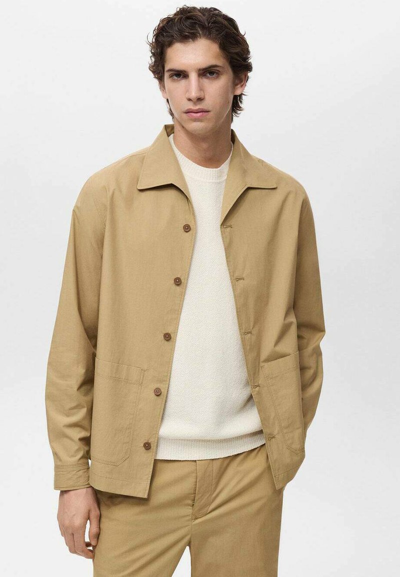 Beige leichte Jacke mit klassischem Kragen, Knopfverschluss und zwei Fronttaschen. Über einem weißen strukturierten Pullover getragen.