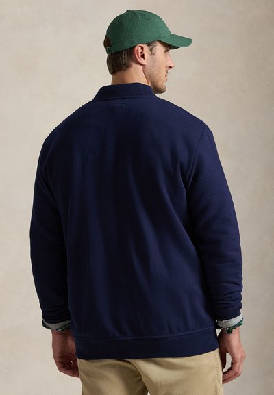 Άνδρας που φοράει μπλε σκούρο πουκάμισο με μακριά μανίκια, χακί παντελόνι και πράσινο καπέλο μπέιζμπολ, με την πλάτη προς το μέρος μας, σε απλό φόντο.