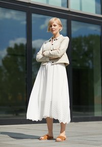 Abito bianco a plissé con orlo a zigzag, abbinato a una giacca leggera beige testurizzata. Indossato con sandali a ciabatta marroni. Ambientazione urbana moderna.