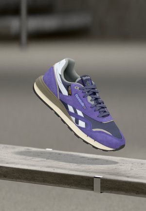 Zapatilla Reebok morada y gris con rayas blancas flotando sobre un riel metálico contra un fondo desenfocado.