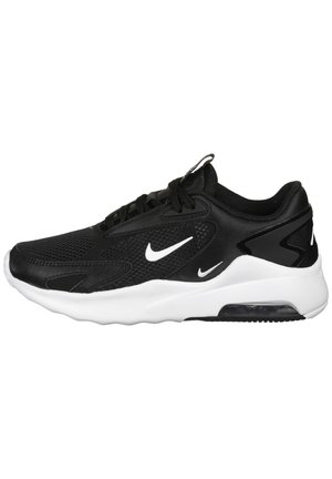 Zapatilla deportiva negra con parte superior de malla y sintético, acentos blancos, suela gruesa y unidad de amortiguación visible. Presenta un diseño de cordones y logo.