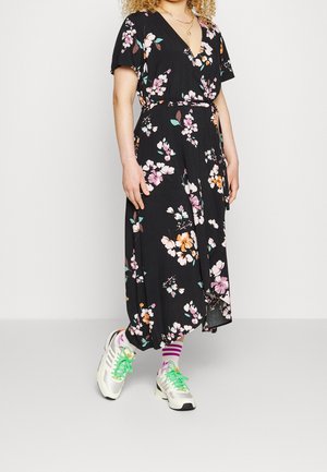 Kvinde iført sort blomsterprint slå-om-kjole med korte ærmer, hvide sneakers med grønne snørebånd og hvide sokker med lilla striber.