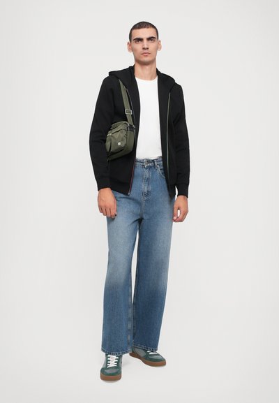 Čierna mikina na zips, biela tričko, široké modré denimové džínsy, zelená crossbody taška a zelené tenisky s hnedými podrážkami.