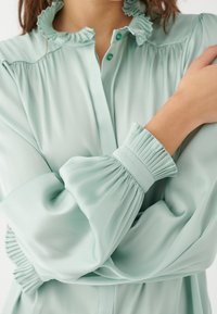 Mintgrünes Bluse mit Rüschenausschnitt und gefältelten Manschetten. Hergestellt aus glattem Stoff, mit Knopfverschluss und lockerer Passform.