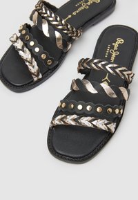 Sandales plates noires avec des brides tressées métalliques et des détails en studs dorés sur un fond blanc.