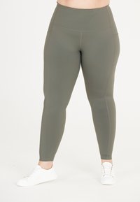 Högmidjade, olivgröna leggings i mjukt, sträckt material med sidfickor och kontrasterande sömmar. Bärs med vita sneakers.