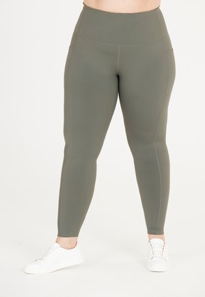 Högmidjade, olivgröna leggings i mjukt, sträckt material med sidfickor och kontrasterande sömmar. Bärs med vita sneakers.