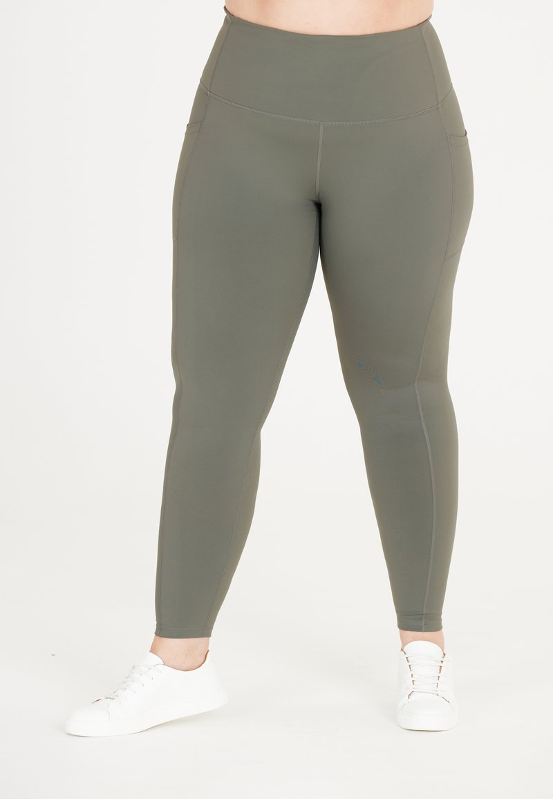 Högmidjade, olivgröna leggings i mjukt, sträckt material med sidfickor och kontrasterande sömmar. Bärs med vita sneakers.
