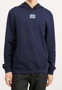 Sweat à capuche bleu marine avec une poche avant, des poignets et un ourlet côtelés. Présente un patch logo carré sur la poitrine. Le matériau semble doux et texturé.