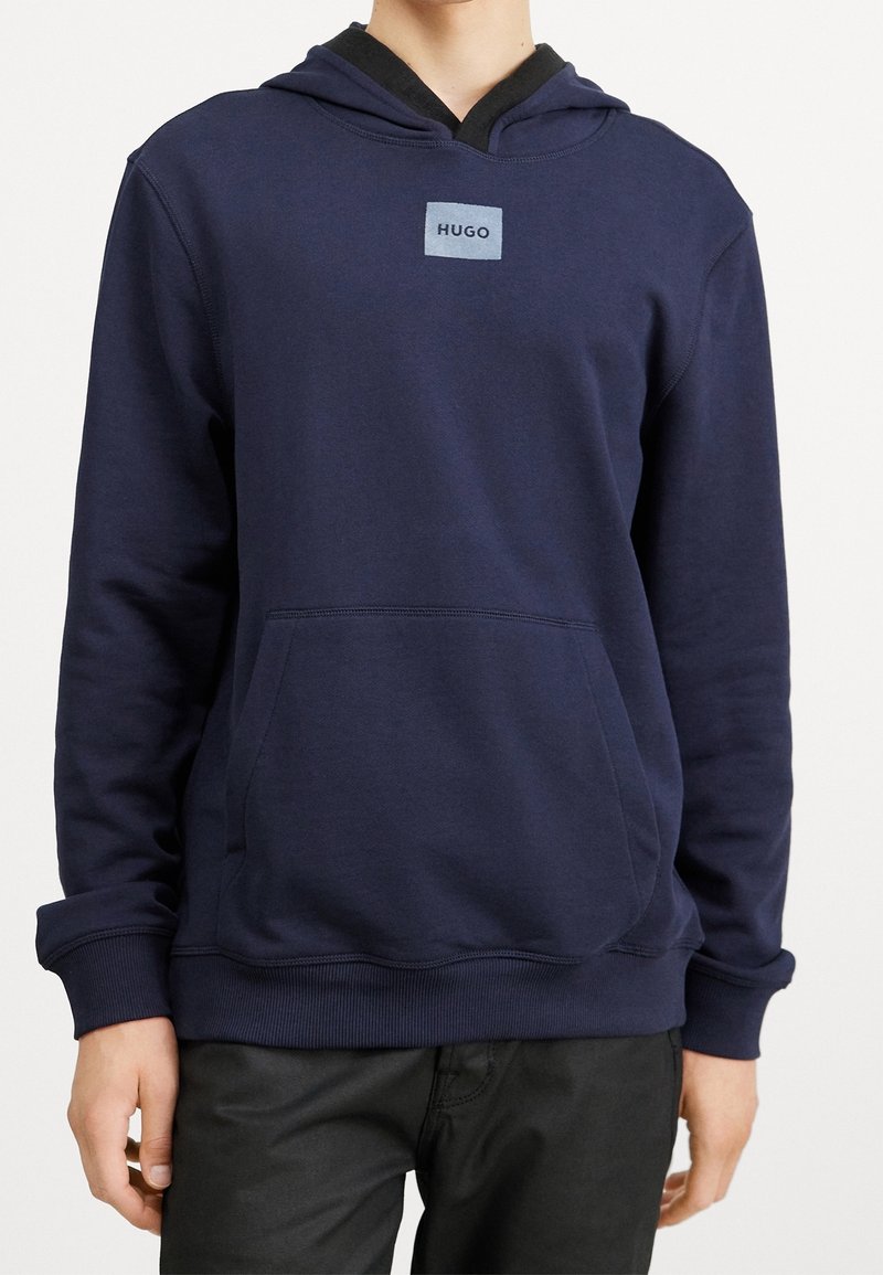 Sweat à capuche bleu marine avec une poche avant, des poignets et un ourlet côtelés. Présente un patch logo carré sur la poitrine. Le matériau semble doux et texturé.