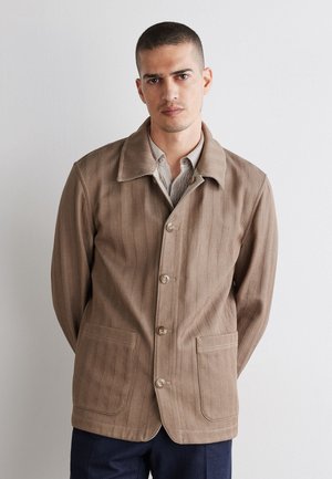 Mann mit kurzen Haaren, der eine beige, geknöpfte Jacke über einem hellen Hemd trägt, steht mit den Händen hinter dem Rücken vor einer weißen Wand.