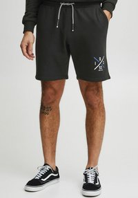 Sorte svedshorts med elastisk talje og snørebånd, med et grafisk print i blå og hvid. Sammen med sorte og hvide sneakers.