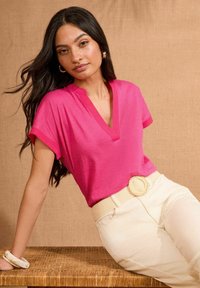 Camiseta fucsia con escote en V y mangas cortas, combinada con pantalones crema y un cinturón trenzado con una gran hebilla redonda, sobre un fondo neutro.
