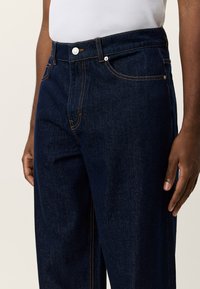 Σκούρο μπλε τζιν με ίσια γραμμή στα πόδια, με αντίθετη ραφή, μπροστινές τσέπες και μεταλλικό κουμπί στη μέση.