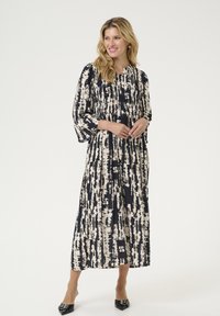 Robe maxi à motif abstrait bleu marine et crème, avec un haut plissé, des manches fluides, une taille cintrée et un tissu doux. Escarpins noirs à talons pointus.