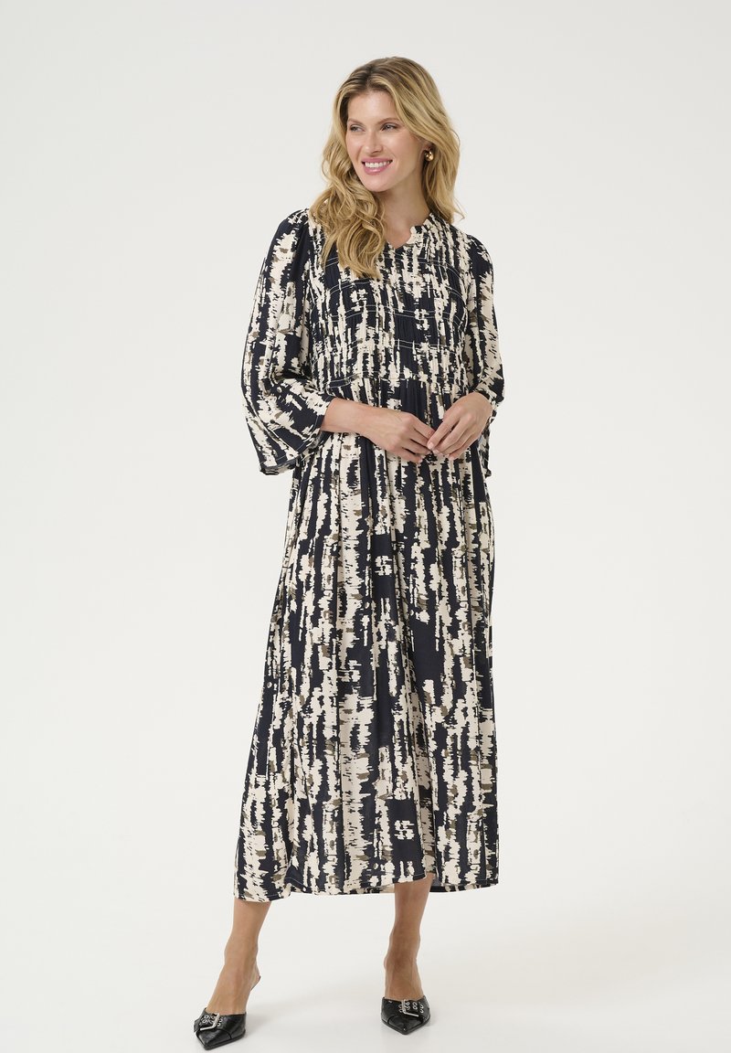 Robe maxi à motif abstrait bleu marine et crème, avec un haut plissé, des manches fluides, une taille cintrée et un tissu doux. Escarpins noirs à talons pointus.