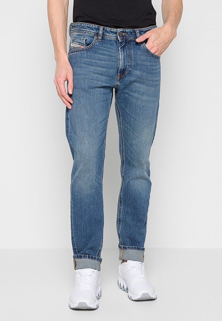 Homme portant un jean roulé bleu moyen, une chemise noire et des baskets blanches, se tenant avec une main dans la poche contre un fond uni.