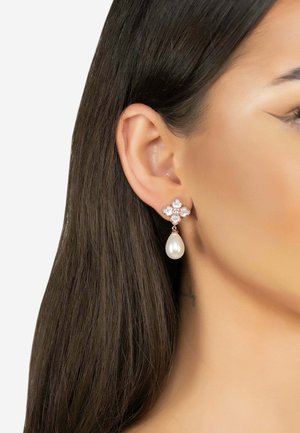 Oreja de mujer con cabello oscuro que lleva pendientes florales de oro rosa con pequeñas perlas y una perla colgante en forma de lágrima.