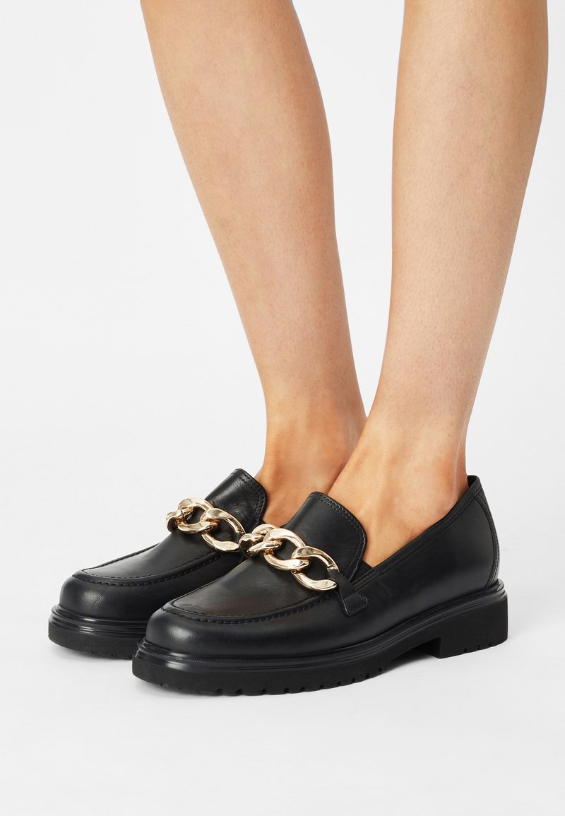 Mocassins en cuir noir avec semelles épaisses portés aux pieds, ornés d'une grande chaîne dorée sur le dessus.