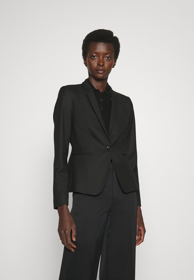 Svart skreddersydd blazer med en knapplukking, notched lapels og sidelommer, iført over en svart skjorte med krage.