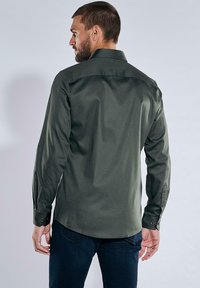 Mann, der sich abwendet, trägt ein dunkelgrünes langärmliges Hemd mit Knöpfen und dunkelblaue Jeans vor einem schlichten hellen Hintergrund.
