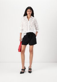 Hvit broderte skjorte med lange ermer, svarte shorts med snor, rød håndveske og svarte høyhelte sko med spenner.