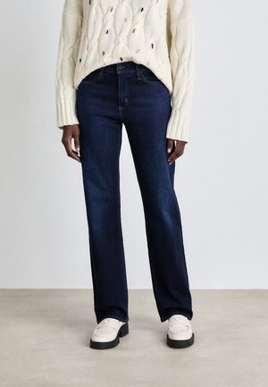 Persoon die donkerblauwe jeans met rechte pijpen, crèmekleurige kabelgebreide trui en witte platform loafers draagt en staat op een neutrale vloer.
