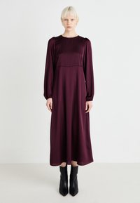 Pitkähihainen, lattiaa myöten ulottuva syvän burgundinpunainen satiinimekko. Ominaisuuksiin kuuluu pyöreä pääntie ja puffihihat. Pari mustia korkokenkiä.