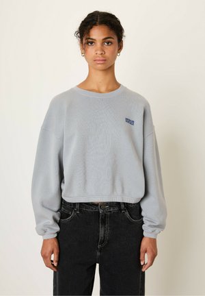 Ljusgrå beskuren sweatshirt med långa ärmar och ribbad textur. Har en blå "American Vintage"-logotyp på vänster bröst. Bärs tillsammans med svarta jeans.